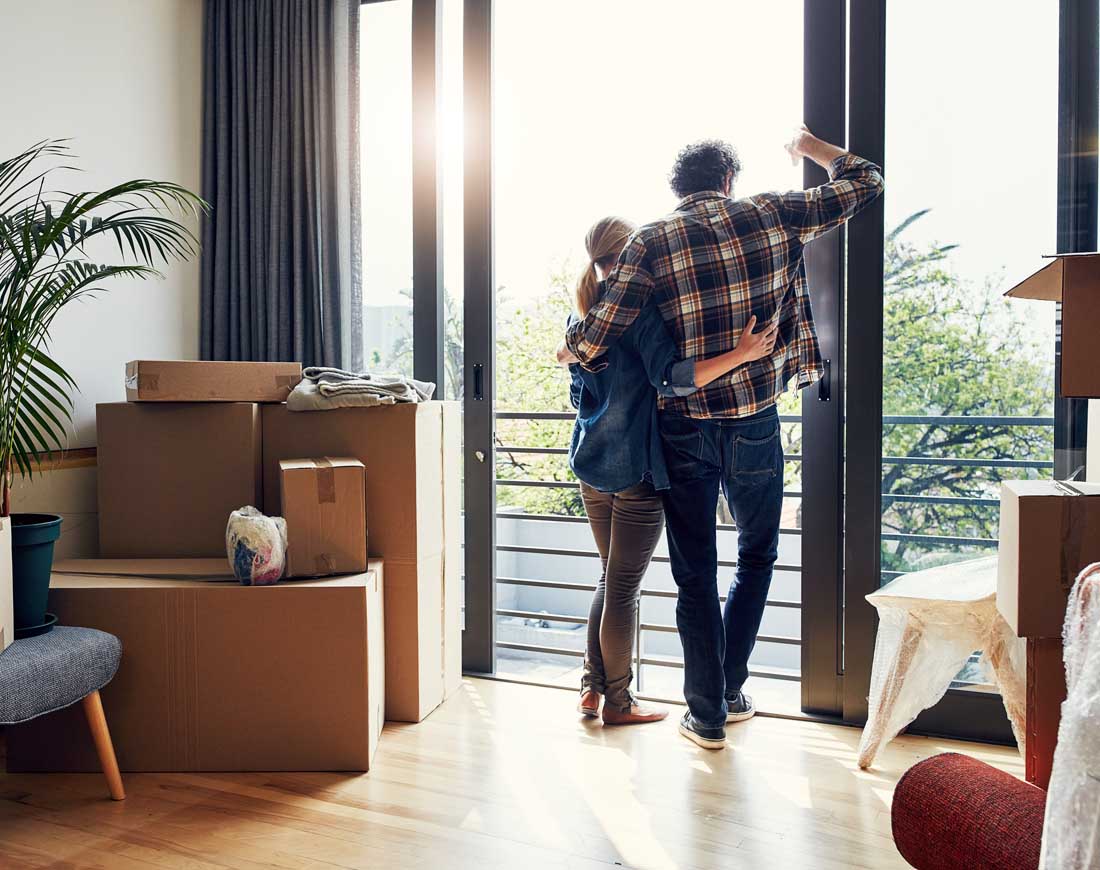 Ein Paar steht Arm in Arm am bodentiefen Fenster der neuen Wohnung und blickt hinaus, in der Wohnung stehen Umzugskartons und verpackte Möbel